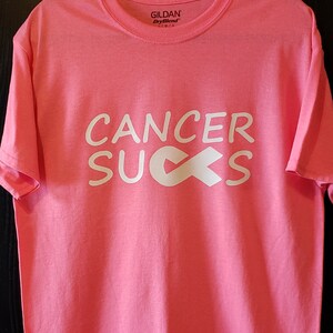 May include: Pink t-shirt with the words "CANCER SUCKS" in white block letters and a white ribbon graphic. The shirt is made of a soft material and has a crew neck. The brand name "Gildan" is visible on the tag.