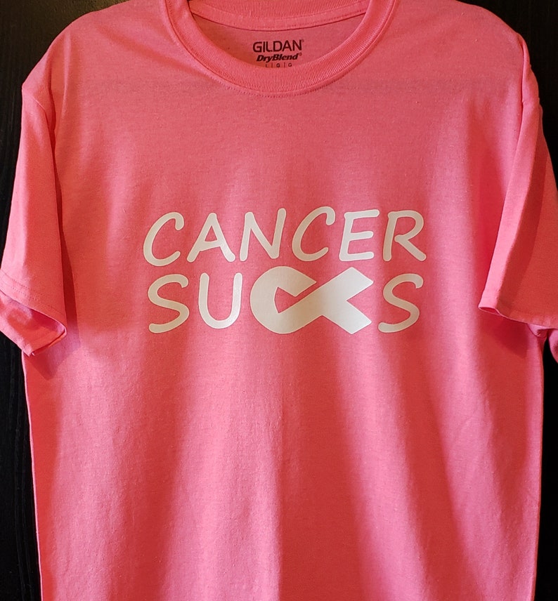 May include: Pink t-shirt with the words "CANCER SUCKS" in white block letters and a white ribbon graphic. The shirt is made of a soft material and has a crew neck. The brand name "Gildan" is visible on the tag.