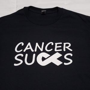 May include: Black t-shirt with the words "CANCER SUCKS" in white, along with a white awareness ribbon. The shirt is made of a soft material and has a crew neck.
