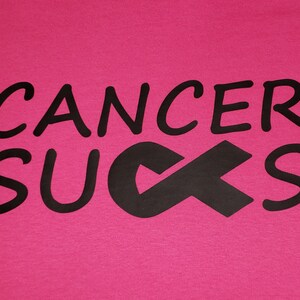May include: A hot pink t-shirt with the words "CANCER SUCKS" in black, along with a black awareness ribbon. The text is in a bold, rounded font.