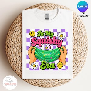 May include: A white t-shirt featuring a graphic print with a purple and white checkered background. The text reads In My Squishy Era in pink and green. The design includes two hands stretching green slime with colorful stars, smiley faces, and flowers.