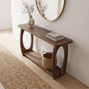 May include: A dark brown wooden console table with a curved design and oval cutouts. A woven basket sits on the lower shelf. A vase with branches and books are on top. A round mirror hangs on the wall.