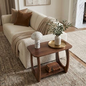 May include: A living room scene featuring a cream-colored sofa with brown velvet pillows and a tan throw blanket. A dark wood side table holds a marble mushroom lamp, a gold tray with a white vase, and a stack of books. A rug and fireplace complete the scene.