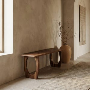 May include: A wooden bench with a rectangular top and curved, open-circle legs. The bench is made of dark brown wood and is placed against a light brown wall. A large, tan vase with branches is next to the bench.