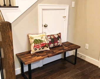 May include: A wooden bench with black metal legs sits under a staircase. Two decorative pillows with holiday-themed patterns are placed on the bench. The floor is made of dark wood.