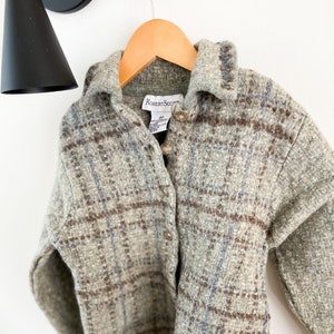 May include: A gray, button-down jacket with a plaid pattern in brown and blue. The jacket is hanging on a wooden hanger. The label reads "Robert Scott".