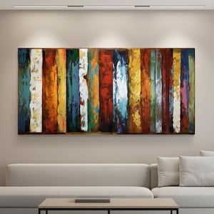 Puede incluir: Obra de arte abstracta con bandas verticales de color. La obra presenta una variedad de tonos, incluyendo blanco, naranja, amarillo, azul y rojo. La pintura está montada en una pared sobre un sofá de color claro.