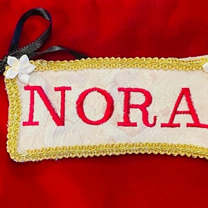 May include: A white fabric banner with gold trim and the name "NORA" embroidered in red. The banner is decorated with two white flowers.
