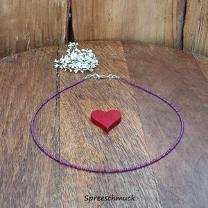 Könnte beinhalten: Eine zarte, lila Perlenkette mit Silberverschluss, ein kleines rotes Herz und weiße Blumen auf einer Holzoberfläche. Der Text "Spreeschmuck" ist unten sichtbar.