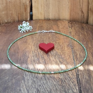 Könnte beinhalten: Eine zarte grüne Perlenkette mit Silberverschluss. Die Halskette wird auf einer Holzoberfläche neben einem kleinen roten Herz und weißen Blumen präsentiert. Die Halskette ist etwa 35,5 cm lang.