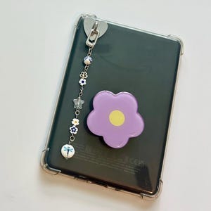 May include: A clear phone case with a decorative charm and a purple flower-shaped phone grip. The charm features a silver heart-shaped clip, blue and white beads, stars, and a dragonfly bead.