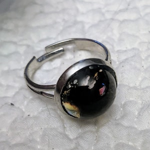 May include: A silver adjustable ring with a black cabochon stone set in a silver bezel. The stone has a speckled pattern of white, gold, and pink.