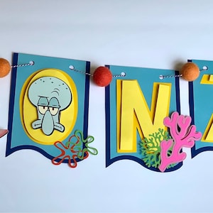 May include: A colorful banner with the name "MONZIE" spelled out in yellow letters. Each letter is decorated with a different character from the cartoon Spongebob Squarepants. The banner is strung with colorful pom-poms.
