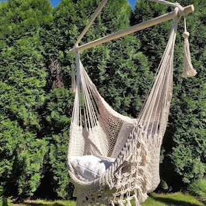 May include: A white macrame hammock chair with fringe hanging from a wooden frame. The hammock is outdoors in a grassy area with green trees in the background.
