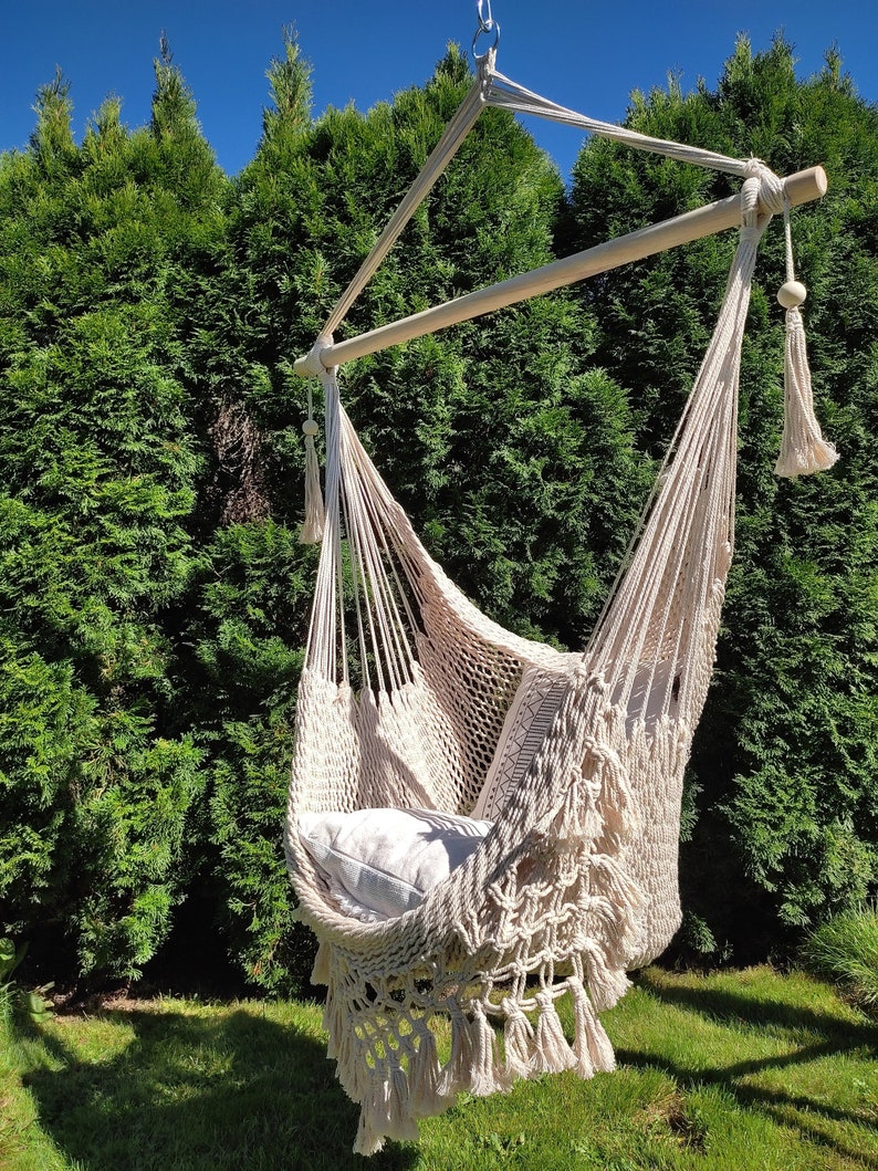 May include: A white macrame hammock chair with fringe hanging from a wooden frame. The hammock is outdoors in a grassy area with green trees in the background.
