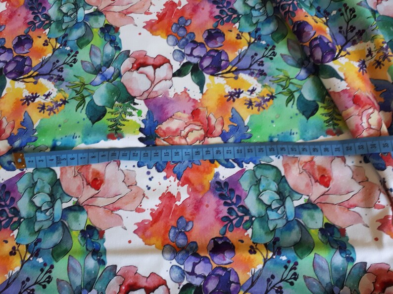 Peut inclure: Tissu floral aquarelle sur fond blanc. Le tissu pr&eacute;sente un motif color&eacute; de fleurs et de feuilles roses, violettes, bleues, vertes et jaunes. Un m&egrave;tre ruban est plac&eacute; sur le tissu pour montrer l'&eacute;chelle.