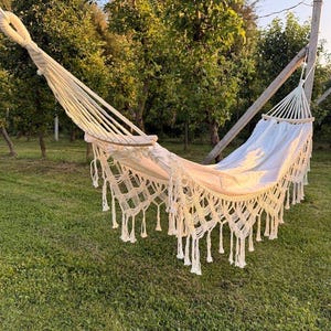 Puede incluir: Una hamaca de color crema con intrincados detalles de flecos de macramé a lo largo de los bordes. La hamaca está suspendida por gruesas cuerdas tejidas unidas a barras separadoras de madera en ambos extremos, creando una forma curva profunda.