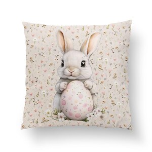 Pode incluir: Um coelho branco segurando um ovo de P&aacute;scoa rosa e branco com bolinhas. O coelho est&aacute; sentado em um fundo bege com um padr&atilde;o floral.
