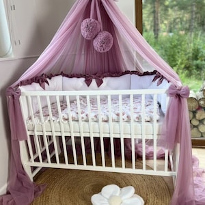 May include: A white cot with a mauve canopy and matching bows. The cot has a floral patterned blanket and white pillows. Two mauve pom-poms hang from the canopy. A white flower-shaped pillow rests on a round, woven rug.