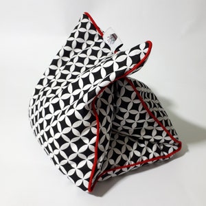 May include: A black and white geometric patterned pillow with red piping. The pillow is folded in half and is sitting on a white surface.