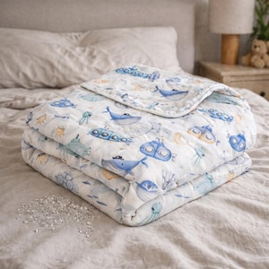 Könnte beinhalten: Eine gefaltete weiße Babydecke mit einem skurrilen Unterwasser-Thema. Die Decke zeigt blaue Wale, U-Boote und andere Meeresbewohner in Blau-, Orange- und Grüntönen. Die Decke ist aus einem weichen, gesteppten Material gefertigt.