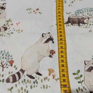 May include: White fabric with a repeating pattern of raccoons, mushrooms, and trees. The raccoons are brown and white with black eyes and noses. The mushrooms are red and white. The trees are green with brown trunks.