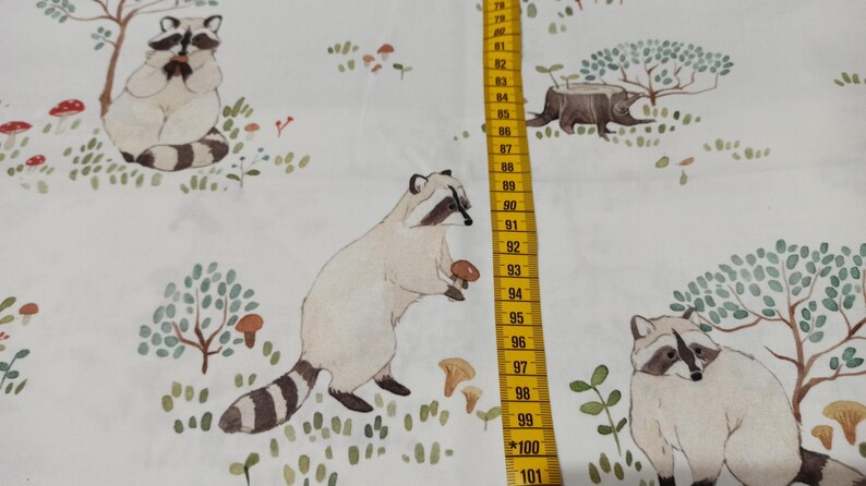 May include: White fabric with a repeating pattern of raccoons, mushrooms, and trees. The raccoons are brown and white with black eyes and noses. The mushrooms are red and white. The trees are green with brown trunks.