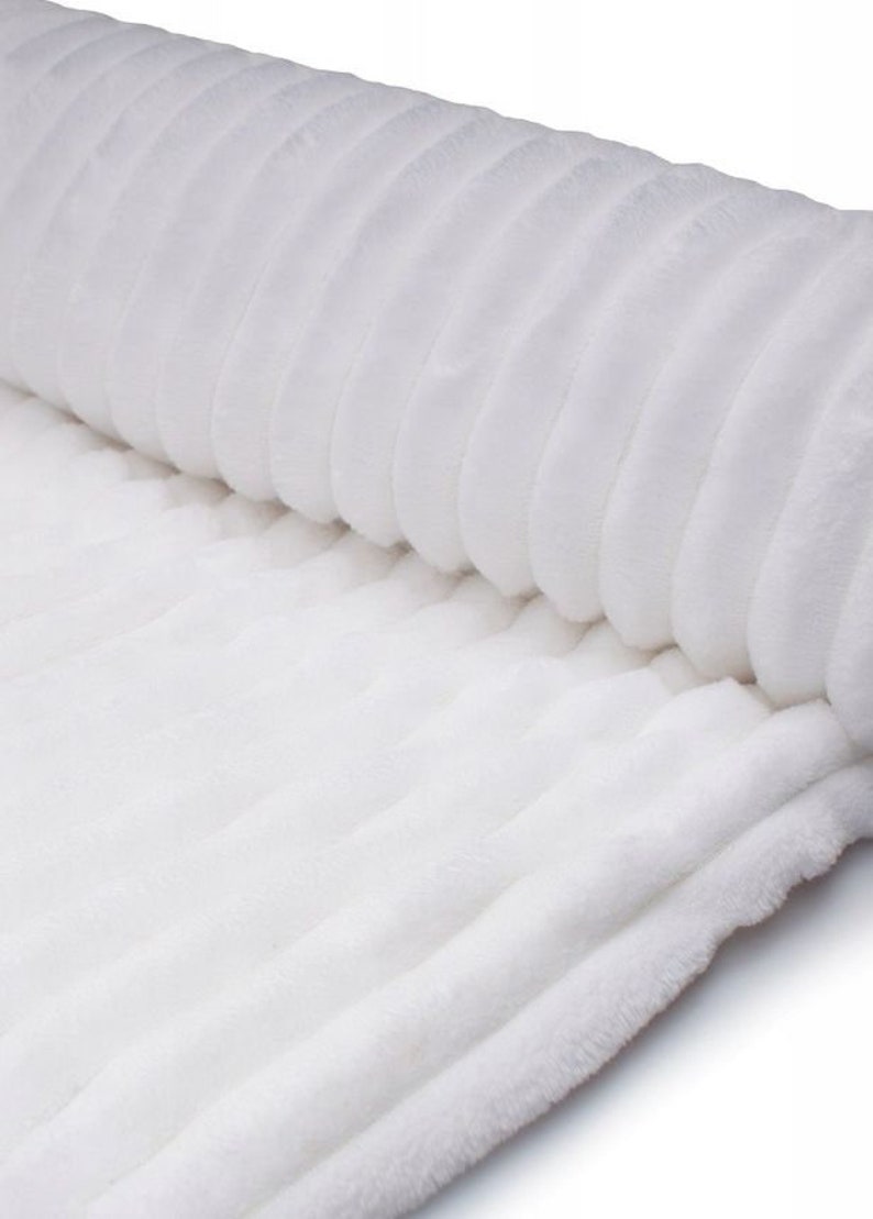 May include: A rolled-up, white, plush fabric with a ribbed texture. The soft material is a solid white color, and the ridges run parallel to each other, creating a pattern. The fabric appears to be a blanket or throw.