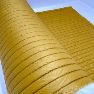 Può includere: Un rotolo di tessuto trapuntato giallo senape con cuciture orizzontali. Il tessuto è arrotolato e il retro bianco è visibile.