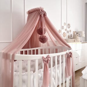 Puede incluir: Una cuna ovalada blanca con un dosel de tul rosa y lazos decorativos. El dosel presenta dos pompones y está drapeado sobre la cuna. La cuna tiene un parachoques blanco acolchado.