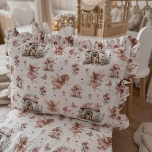 Könnte beinhalten: Ein weißes Kissen- und Deckenset zum Thema Feen. Das Set zeigt ein sich wiederholendes Muster aus Feen, Pilzhäusern, Blumen und Schmetterlingen in Rosa- und Brauntönen. Das Kissen hat einen gerüschten Rand.