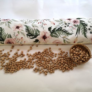 Könnte beinhalten: Ein langes, rechteckiges Kissen mit Blumenmuster, gefüllt mit braunen Bohnen. Das Kissen liegt auf einer weißen Oberfläche, daneben ein kleiner Korb mit braunen Bohnen.
