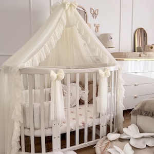 Könnte beinhalten: Ein weißes, ovales Babybett mit Baldachin und Rüschen-Tüll. Das Bett hat vertikale Sprossen und ist mit Schleifen verziert. Die Bettwäsche weist ein Blumenmuster auf. Dekorative Kissen und ein blütenförmiges Bodenkissen sind ebenfalls zu sehen.