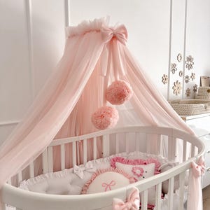 May include: An oval white cot with a pink canopy and matching bedding. The canopy is made of sheer fabric and decorated with bows and pom-poms. The bedding includes pillows with bow details, creating a soft and elegant nursery setting.