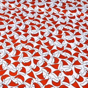 Op de afbeelding: Witte stof met een herhalend patroon van rode kerstmutsen, elk met witte rand en een witte pompon. De mutsen zijn in verschillende richtingen gerangschikt, wat een feestelijk kerstontwerp creëert.