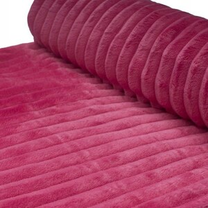 May include: A roll of plush, ribbed fabric in a vibrant magenta hue. The fabric has a soft, velvety texture and a series of parallel ridges running across its surface. The material is partially rolled, revealing the texture and color.