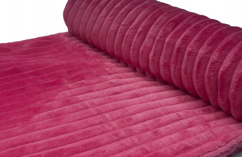 May include: A roll of plush, ribbed fabric in a vibrant magenta hue. The fabric has a soft, velvety texture and a series of parallel ridges running across its surface. The material is partially rolled, revealing the texture and color.