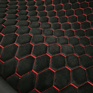 Può includere: Un tessuto nero caratterizzato da un motivo esagonale ripetuto, cucito con filo rosso brillante. La superficie presenta un aspetto morbido e testurizzato, con il design geometrico a nido d'ape che crea una griglia coerente sul tessuto.