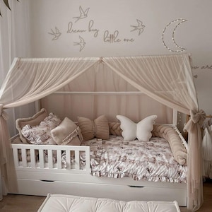 Könnte beinhalten: Ein weißes Kleinkindbett mit einem beigefarbenen Baldachin und einem Bettwäsche-Set mit Blumenmuster. Das Bett hat ein weißes Kopfteil und Fußteil mit einem weißen Geländer. Die Bettwäsche umfasst eine Bettdecke mit Blumenmuster, ein weißes Kissen mit Schmetterlingsflügeln und ein beiges Kissen. Die Wand hinter dem Bett ist mit einem dekorativen Mond und einem Zitat verziert, das "dream big little one" lautet.