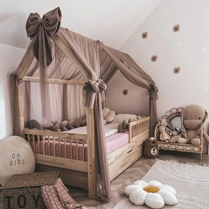May include: A wooden house-shaped bed frame with a sheer, mauve canopy and matching bow accents. The bed is decorated with plush pillows and a pink comforter. A wicker chair with a stuffed bunny and other toys sits nearby.