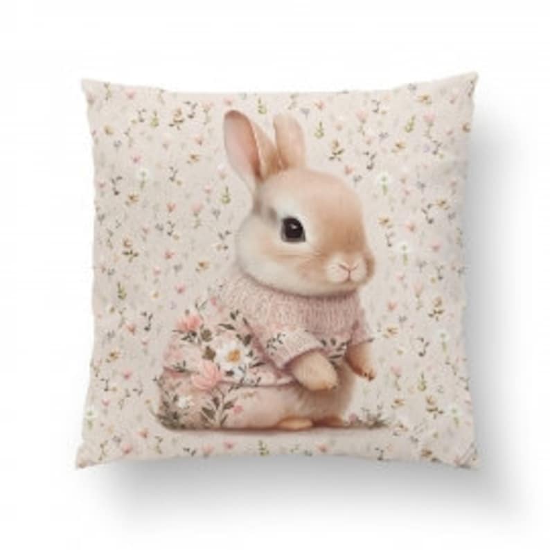 Pode incluir: Uma almofada quadrada de pel&uacute;cia com um coelho marrom claro vestindo um su&eacute;ter rosa com bordados florais. O fundo &eacute; bege claro com um pequeno padr&atilde;o floral.