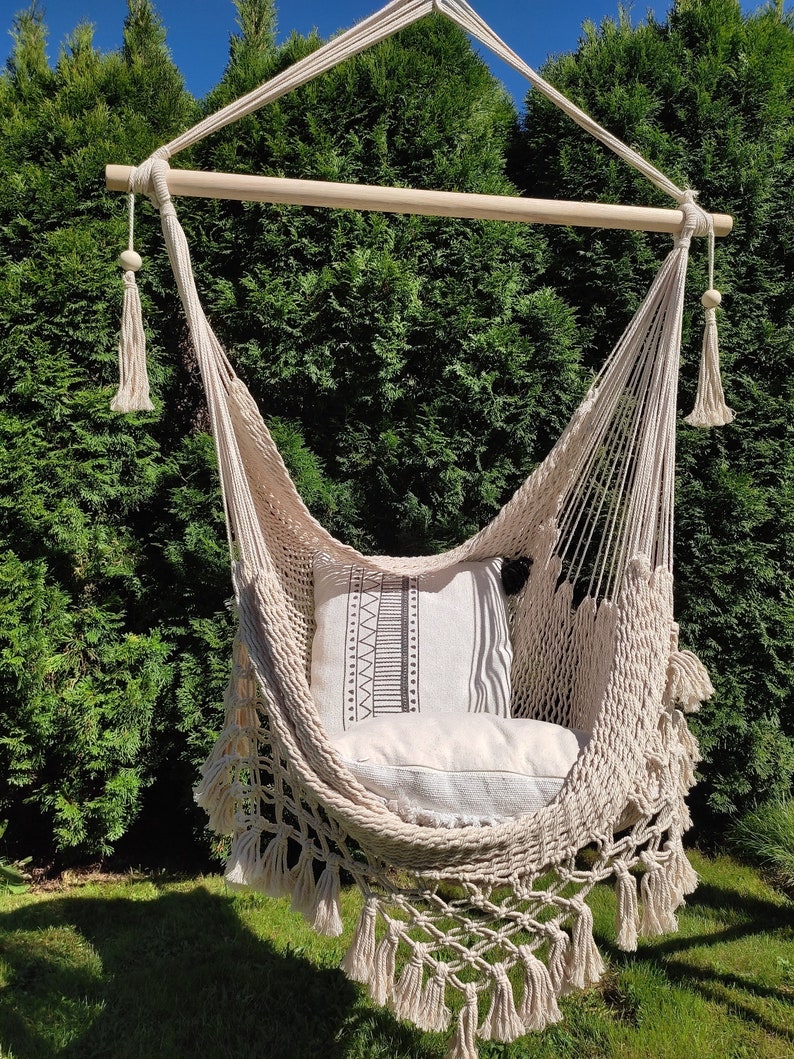 May include: A cream-colored hammock chair with fringe details and a patterned pillow. The hammock is hanging from a wooden bar and is outdoors in a garden setting.