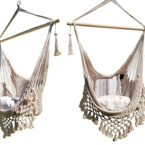 May include: Two beige macrame hanging chairs with fringe details and wooden bars. Each chair has a white cushion with a brown and white patterned pillow.