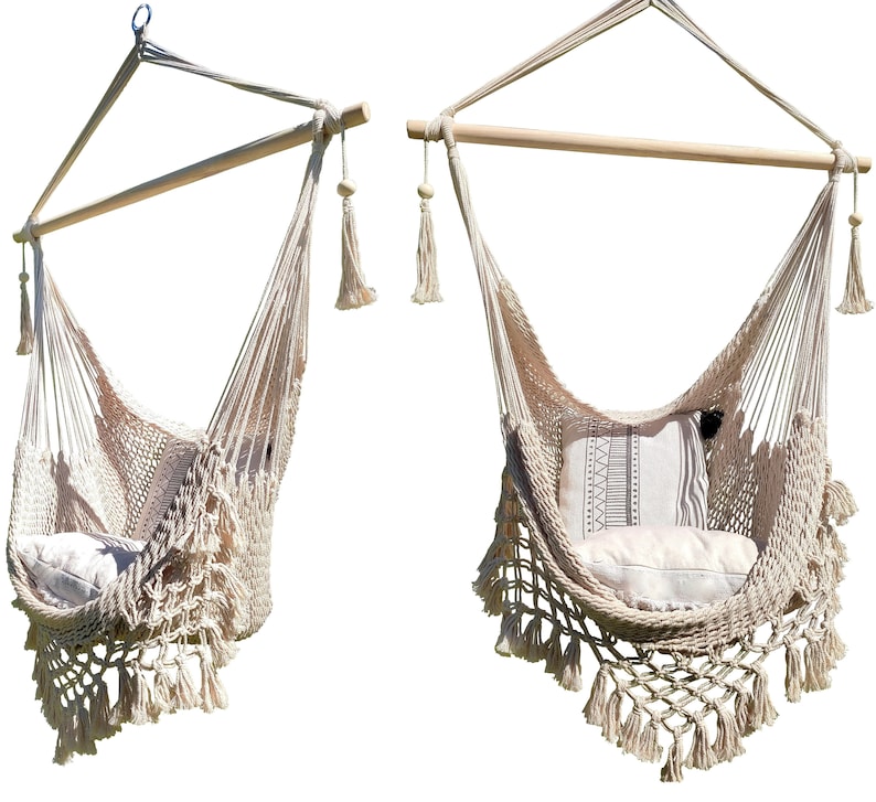 May include: Two beige macrame hanging chairs with fringe details and wooden bars. Each chair has a white cushion with a brown and white patterned pillow.