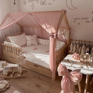 Könnte beinhalten: Ein Holzbettgestell mit einem rosa Baldachin und weißen Bettwäsche. Das Bett hat ein weißes, flauschiges Kissen und ein weißes, geflochtenes Kopfteil aus Seil. Das Bett ist mit einer rosa Schleife verziert und die Worte "dream big little one" sind an der Wand hinter dem Bett geschrieben.