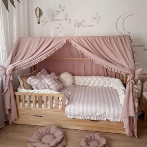 Könnte beinhalten: Ein helles Holzbett mit Baldachin, rosa Bettwäsche und einem passenden Baldachin. Das Bett hat eine gestreifte Bettdecke, dekorative Kissen und zwei blumenförmige Bodenkissen. An der Wand sind ein Heißluftballon, Vögel und der Text "dream big little one" zu sehen.