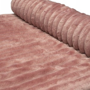 May include: A rolled-up, plush, dusty rose-colored fabric with a ribbed texture. The soft material is partially rolled, revealing the texture and color of the fabric. The fabric appears to be a blanket or rug.