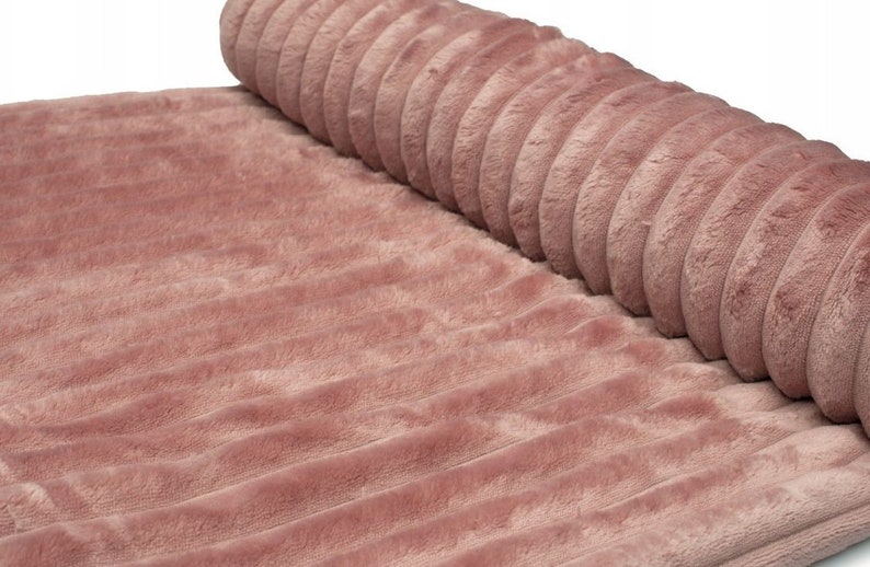 May include: A rolled-up, plush, dusty rose-colored fabric with a ribbed texture. The soft material is partially rolled, revealing the texture and color of the fabric. The fabric appears to be a blanket or rug.