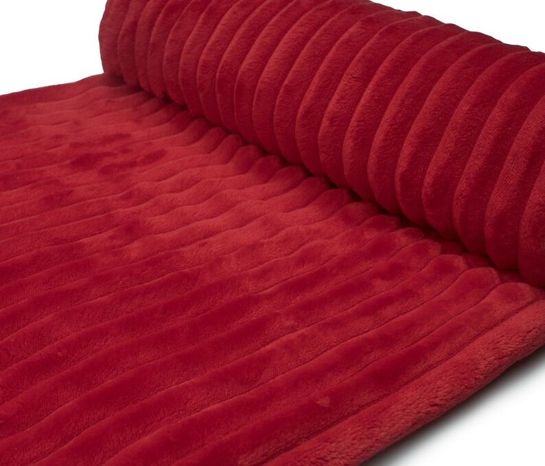 May include: A rolled-up, plush red fabric with a ribbed texture. The fabric is a solid, vibrant red color and appears soft and inviting. The ribbed pattern runs lengthwise across the material, creating a visually interesting surface.