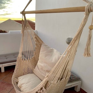 May include: A beige hammock chair with a thick cushion, hanging from a wooden bar. The hammock is made of woven rope and has a fringe detail.